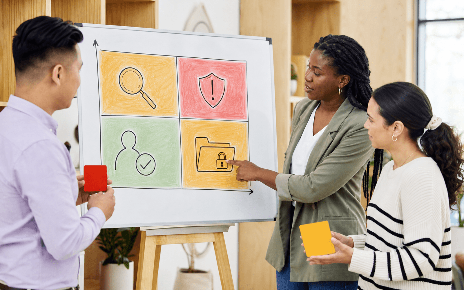A Black woman educator points to an illustrated risk matrix while an East Asian man holds a red tile and a light-skinned woman holds an amber tile in a bright workshop