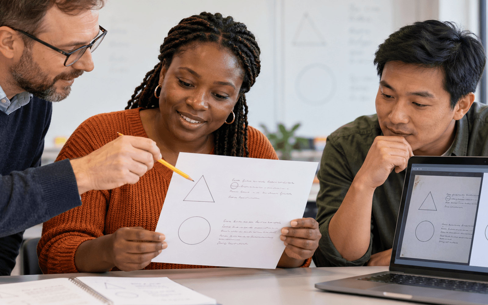 A White man teacher and Black and East Asian adult learners compare a scanned geometry worksheet with a transcription on a laptop.