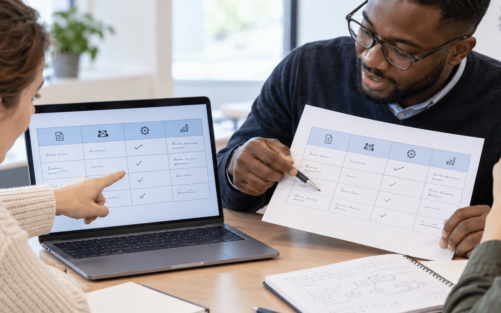 A Black man teacher and adult learners compare a printed table with its reconstructed layout on a laptop.